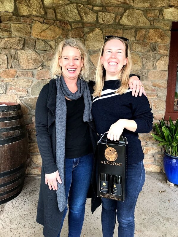 sandy-of-alkoomi-winery-with-her-arm-around-naomi-of-the-travelling-corkscrew-while-holding-a-box-with-a-bottle-of-melaleuca-and-victrix-white-wine-in-frankland-river-great-southern
